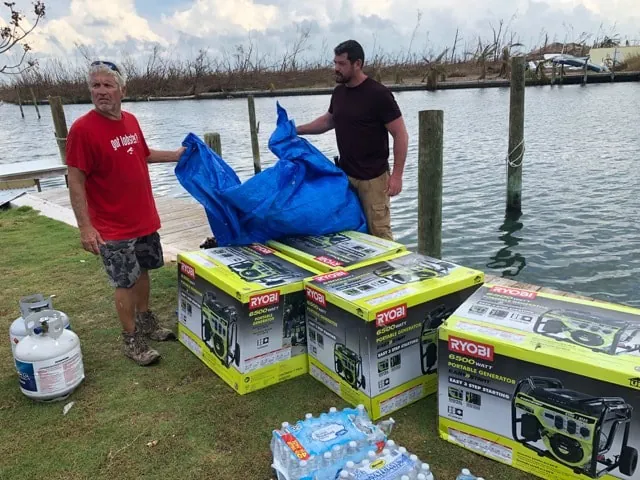 Two men pull a blue tarp off a load of three boxed portable generators, a propane tank and cases of water on a waterfront lawn.
