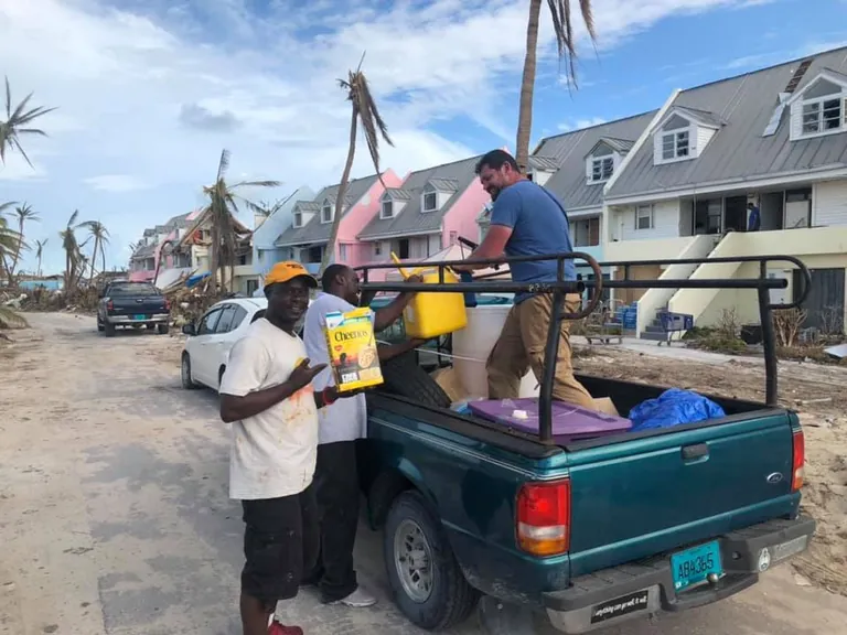 A volunteer in the bed of a green pickup hands a yellow water jug down to two residents, one holding a box of cereal, on a street of storm damaged townhouses.