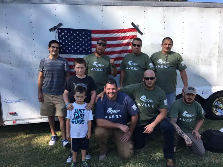 AVERT volunteers in green and navy AVERT shirts, with two boys, in front of a white supply trailer hung with an American flag.